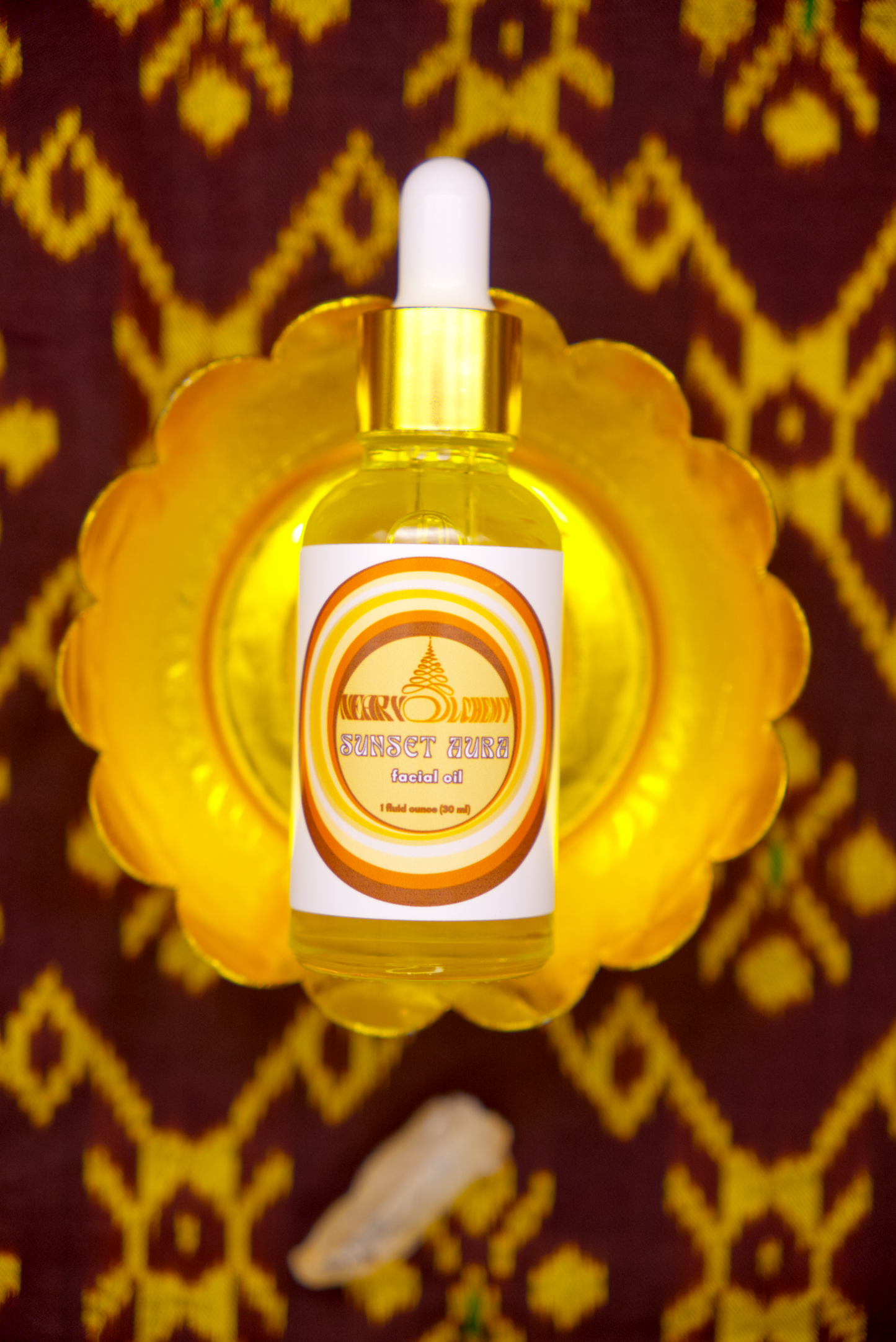 Bottle of Sunset Aura Facial Oil on decorative stand with Khmer silk fabric background and Sunset Aura crystal.