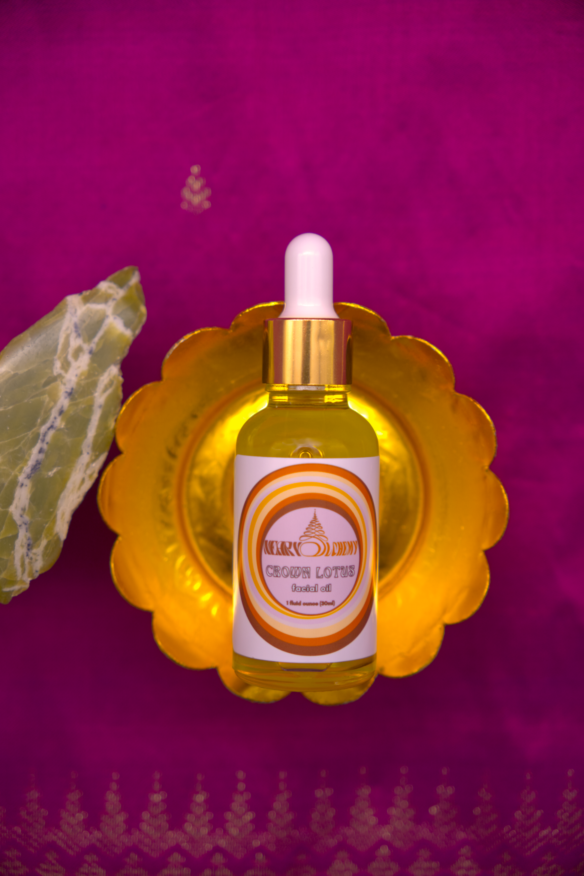 A bottle of Crown Lotus Facial Oil  placed on a decorative container, against a purple Khmer silk fabric with Serpentine crystal.