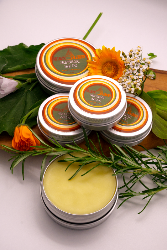 Stack of Supreme Salve containers with with assortment of herbs on wooden plank.