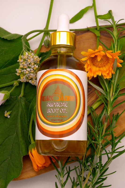 Bottle of Supreme Body Oil surrounded by rosemary, calendula, yarrow, plantain and marshmallow on a wooden plank.