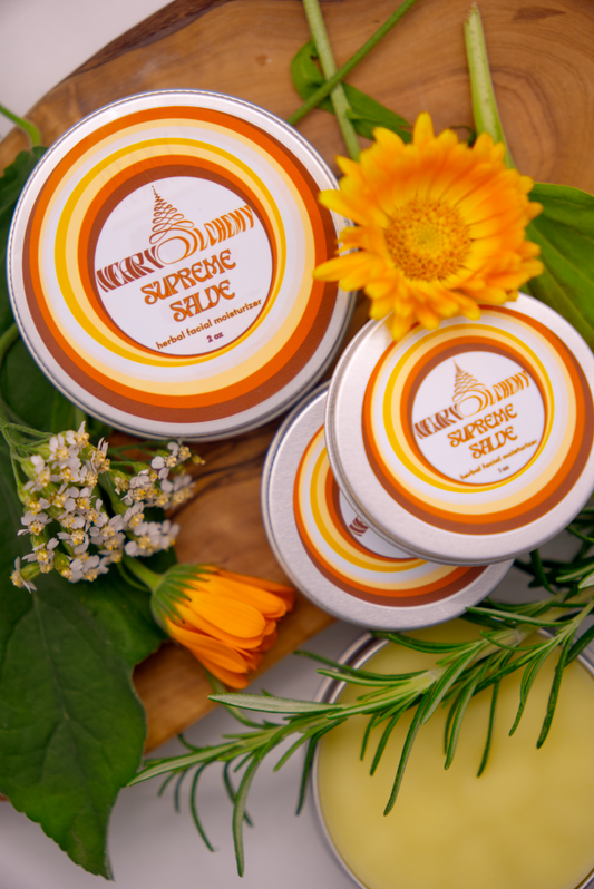 Four tins of Supreme Facial Salve surrounded by various herbs on a wooden plank.