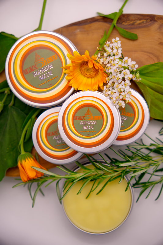 Four containers of Supreme Salve with a decorative arrangement of flowers and herbs on a wooden board.