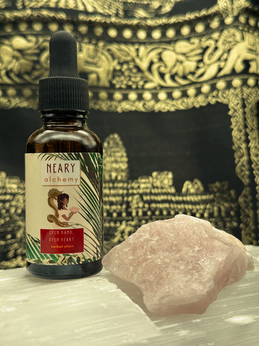 An amber colored tincture bottle with the label "Open Hand Open Heart Elixir" placed next to a rose quartz crystal.